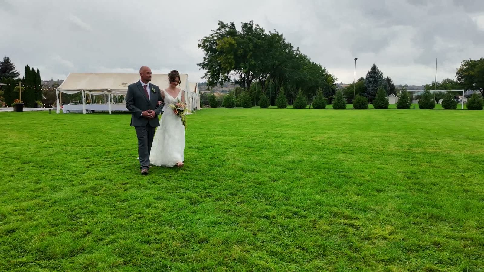 A father walks his daughter, in her wedding dress, across a green field toward the ceremony