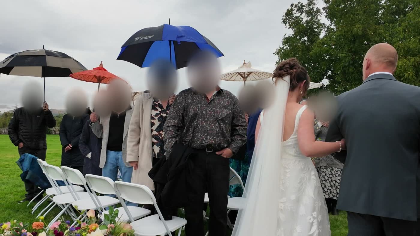 A bride and her father walk down a grassy aisle past guests holding umbrellas, a blue umbrella above them